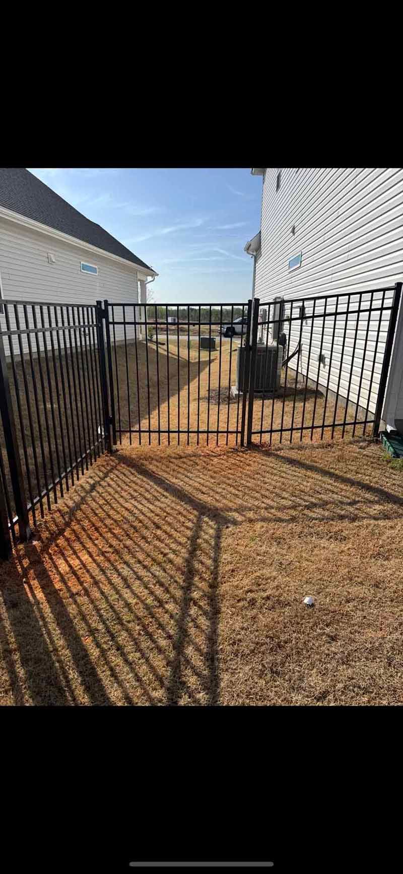 Black metal gate opening to a small dirt path between two white-sided buildings under a blue sky