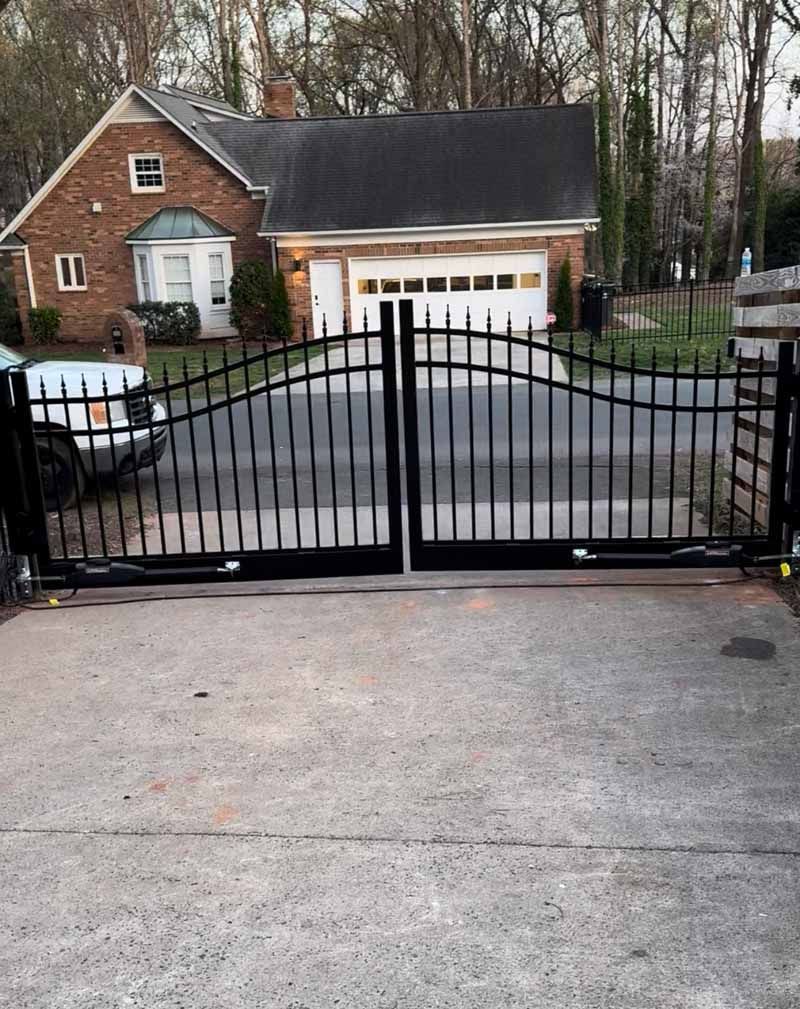 Black metal driveway gate in front of a brick house with a garage and parked cars.