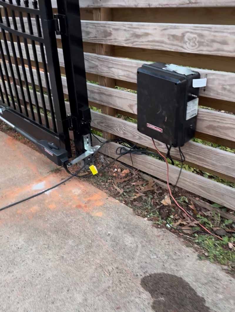Black gate corner with a mounted utility box beside a wooden fence and concrete driveway.