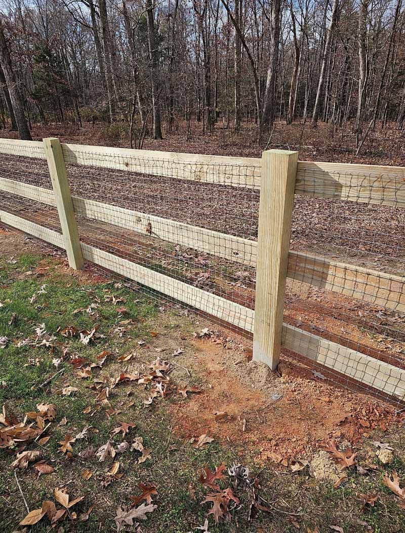 Wooden split-rail fence along a wooded path, with leaf-covered ground and bare trees in the background.