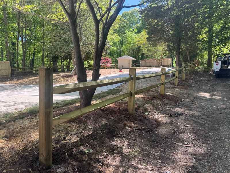 Wooden fence beside a shaded dirt path in a wooded park area with a road beyond.