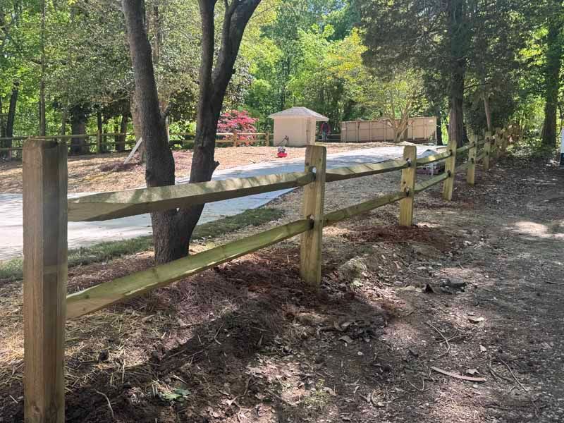 Wooden fence beside a dirt path in a wooded park, with a small tan shelter in the background.