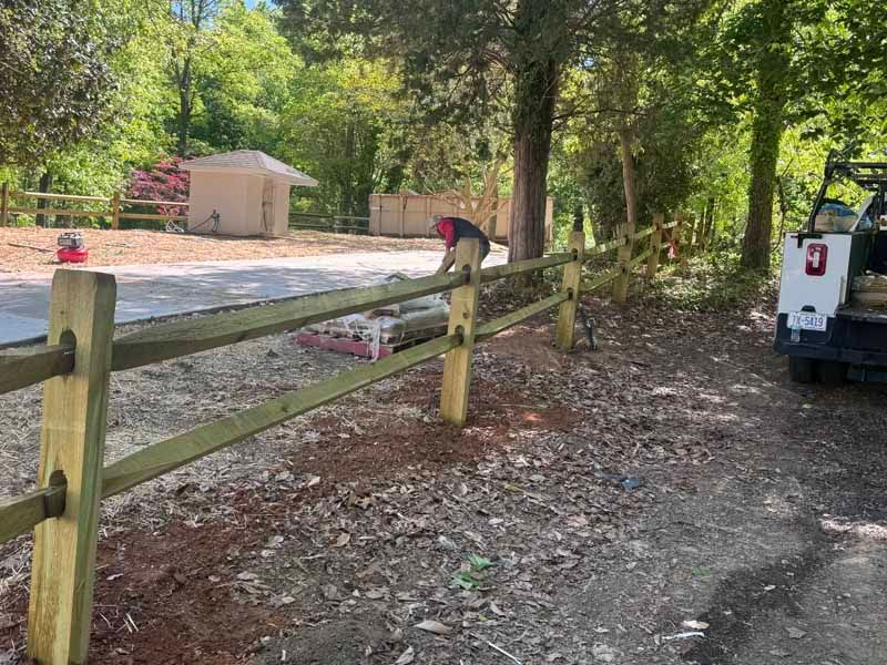 Person in red shirt working beside a wooden fence in a wooded area near a parked truck.