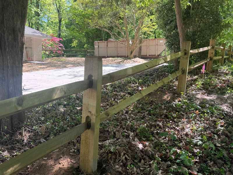 Wooden split-rail fence along a wooded path beside a gravel driveway and gate