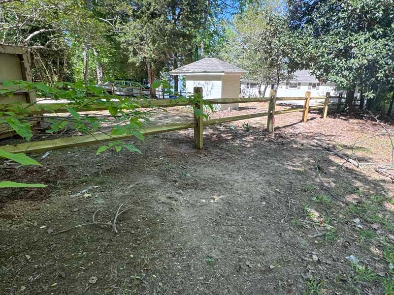Wooden fence along a shaded dirt path beside a small white building and trees