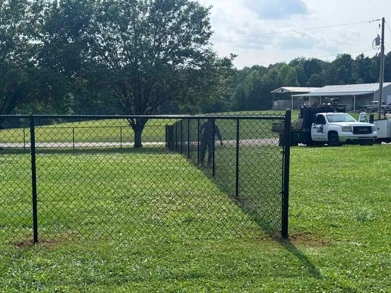 Chain-link fence gate in a grassy yard beside a white pickup truck and house under trees.