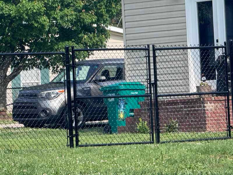 Black SUV parked behind a chain-link fence beside a house and green trash bin.