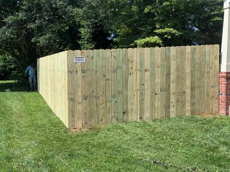 Wooden privacy fence enclosing a grassy yard beside a brick house, with trees in the background.