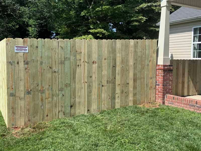Wooden privacy fence beside a house and brick porch, with grass in the foreground.