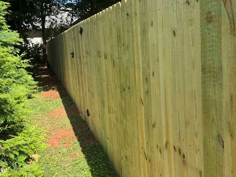Tall wooden privacy fence beside a narrow grassy yard with trees in the background