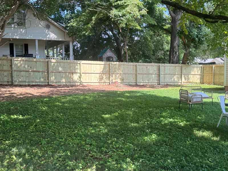 Backyard with green lawn, wooden fence, large trees, and a house porch on the left