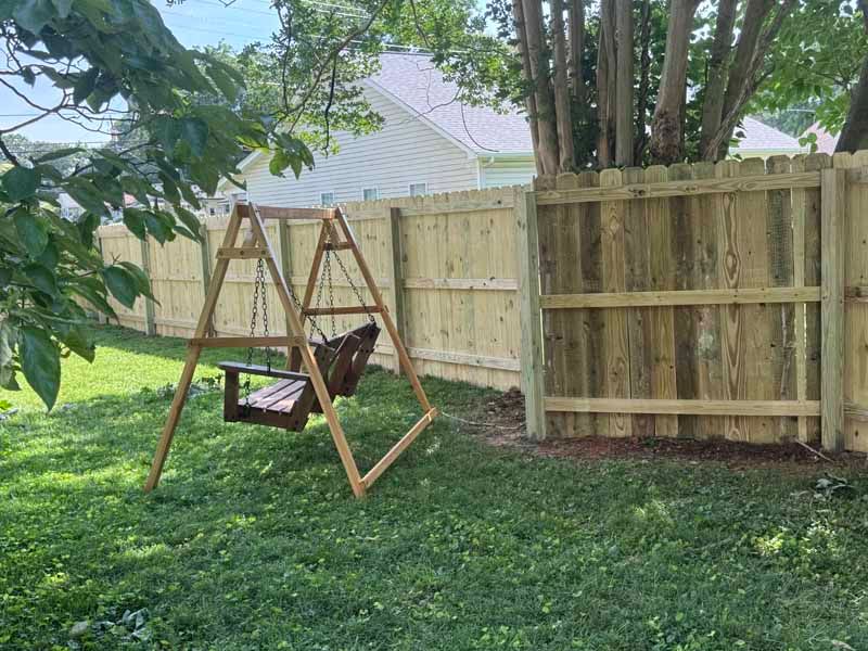 Wooden swing bench under trees beside a fenced backyard lawn