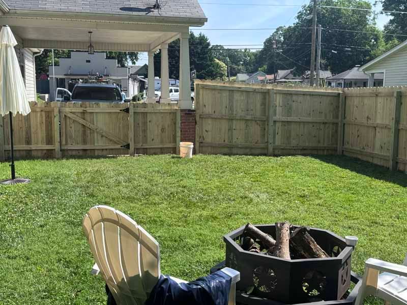 Backyard with green lawn, wooden fence, patio chairs, and a fire pit near a covered porch.
