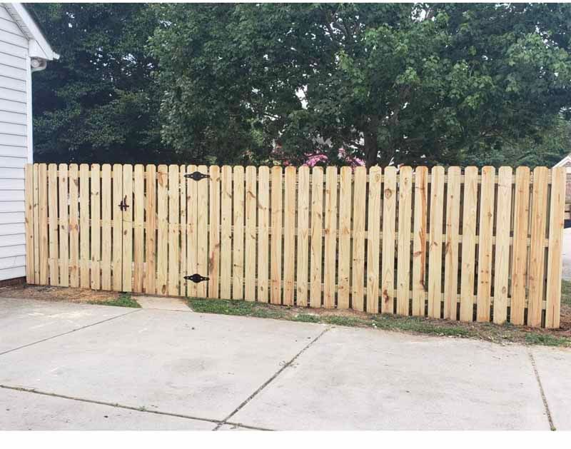 Tan wooden privacy fence beside a concrete driveway, with trees and part of a white house visible.
