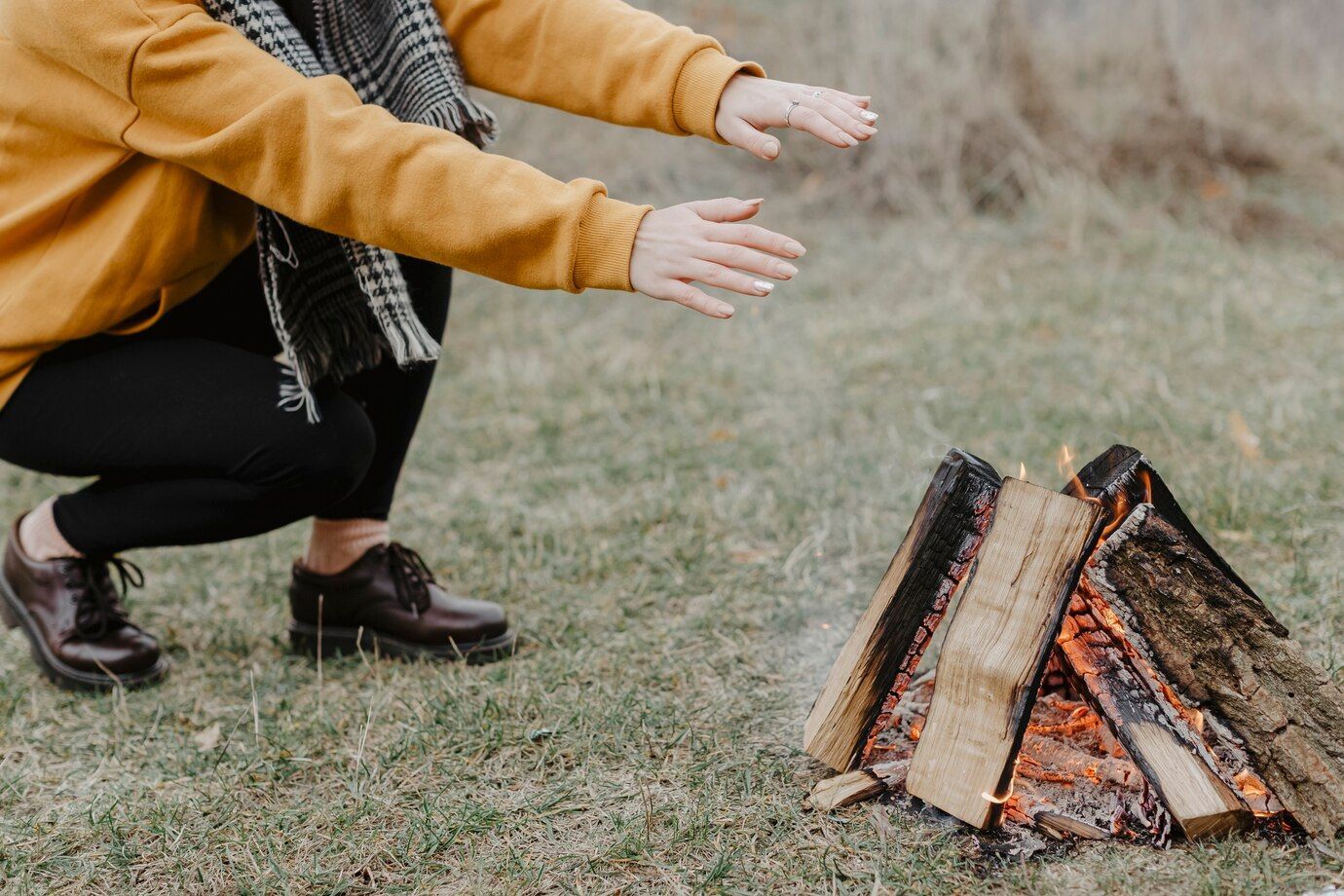 Person warming hands by a campfire outdoors. They wear a mustard yellow sweater and black pants.
