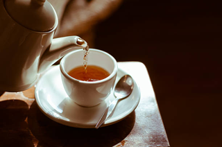 Tea being poured from a white teapot into a white cup on a saucer, with a spoon, on a wooden surface.