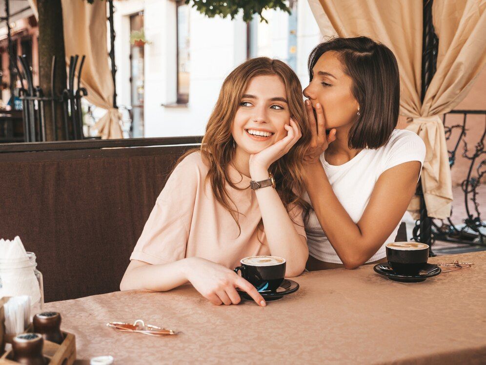 Two women at an outdoor cafe, one whispering to the other who is smiling, coffee cups on the table.