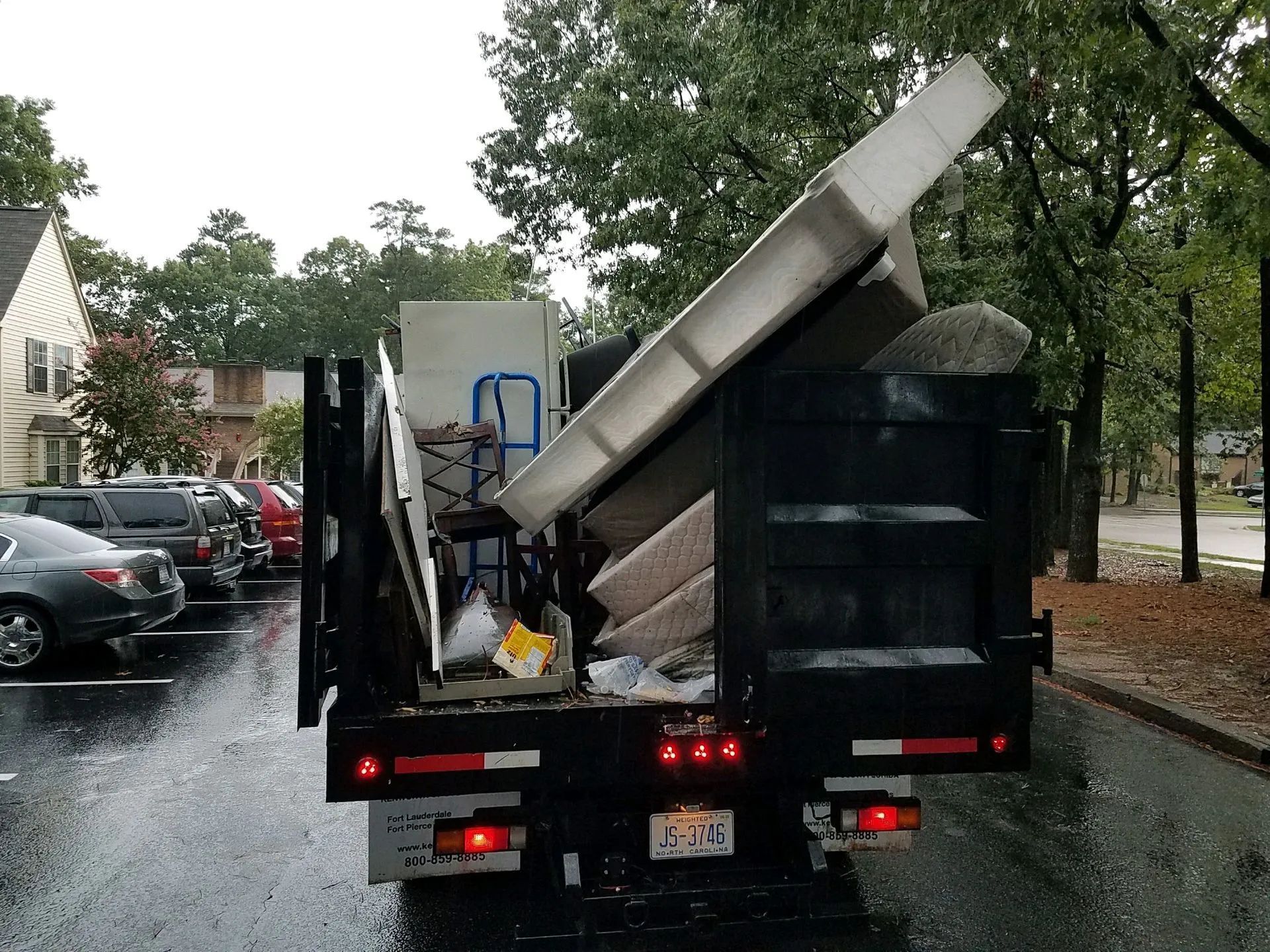 A truck bed overflowing with construction debris, parked in a wet parking lot, near parked cars and trees.