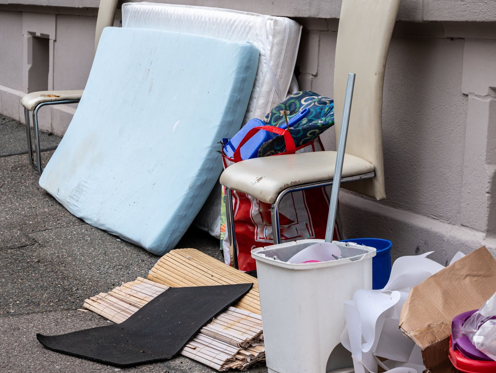 Pile of discarded household items, including mattresses, chairs, and trash bags, on a sidewalk against a building.