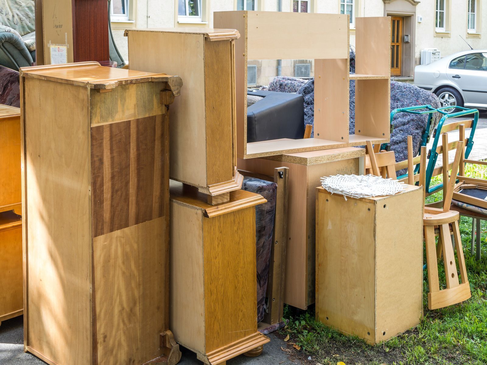Stacked wooden furniture and cabinet pieces outdoors, with a car and house in the background
