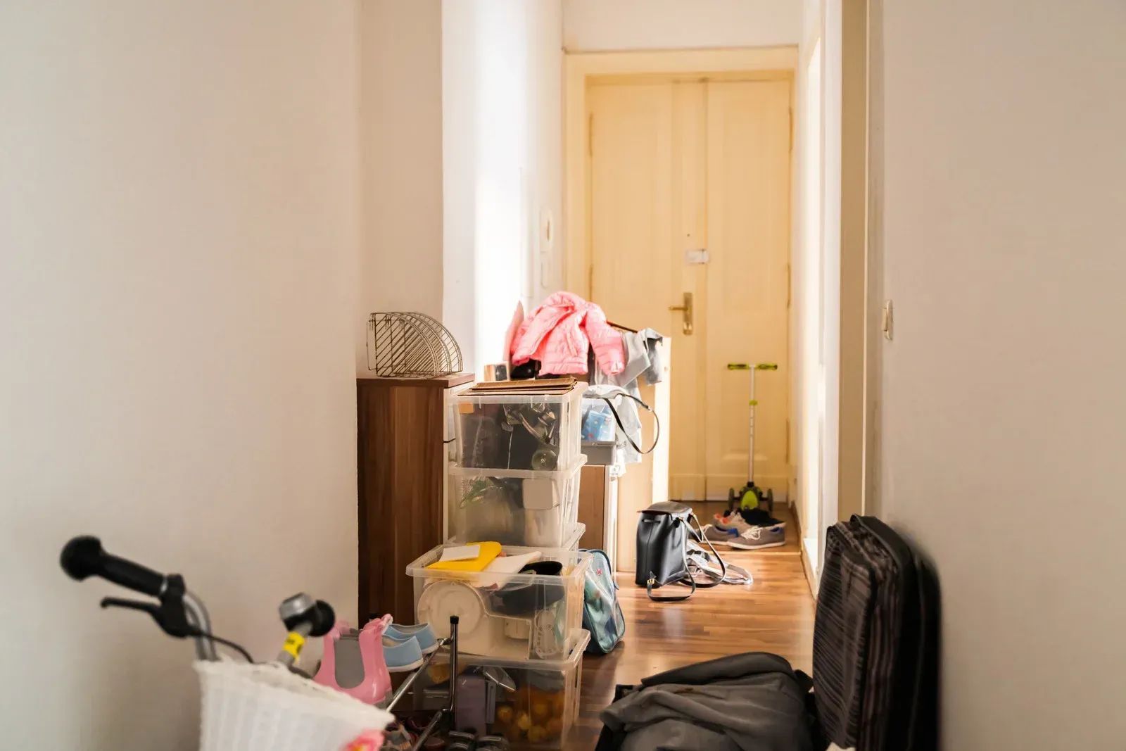 Cluttered hallway with boxes, luggage, clothes, and a bicycle. Door at the end.
