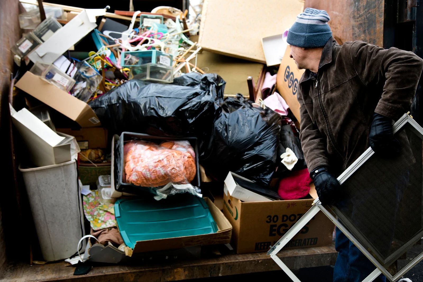 A person sorting through a dumpster filled with trash, holding a window screen.