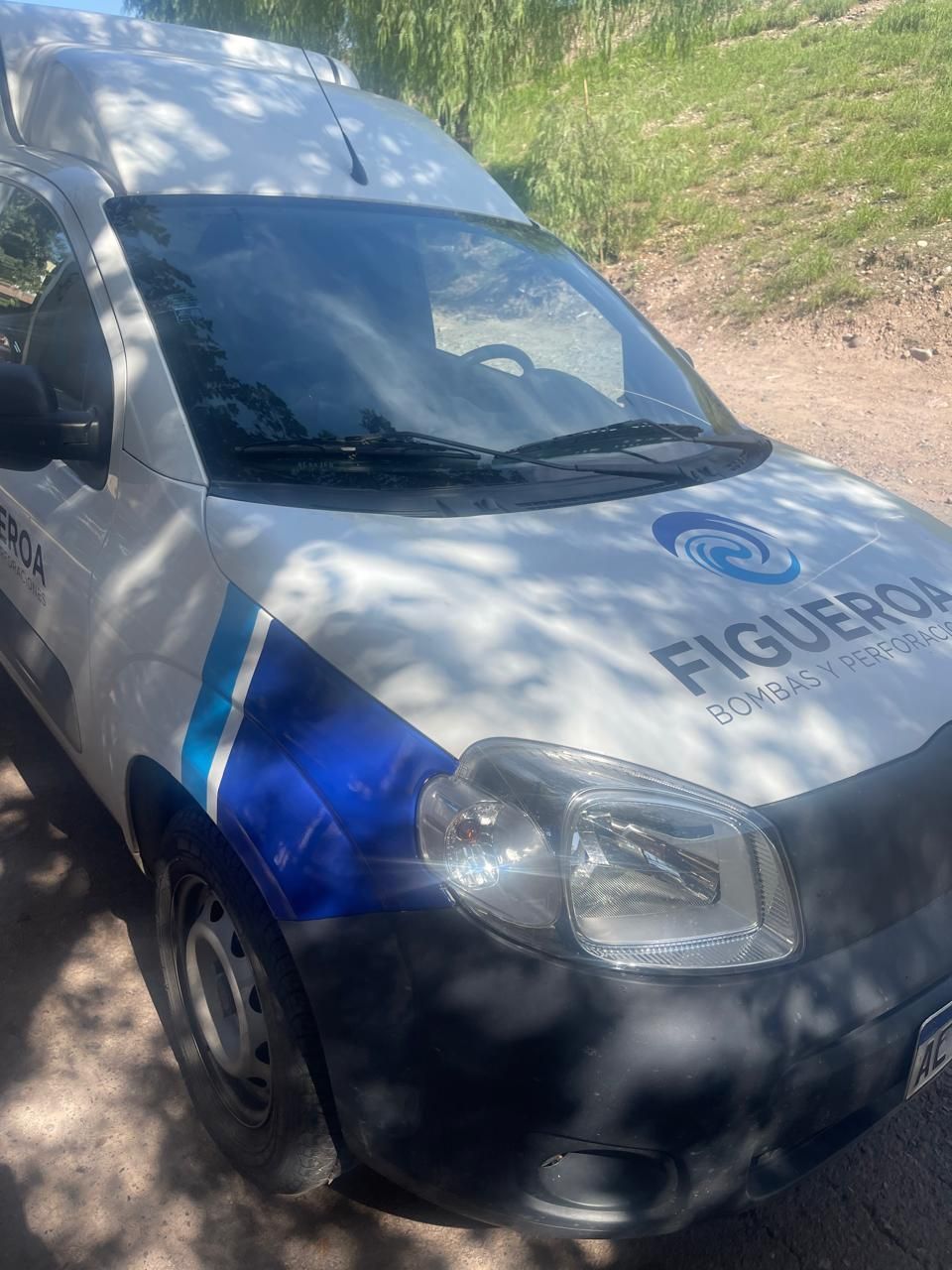 Una furgoneta comercial blanca y azul, a la que le falta una rueda trasera, estaba aparcada en un solar de tierra con árboles al fondo.