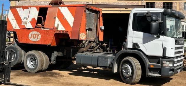 Un camión semirremolque Scania blanco estacionado al aire libre, transportando una gran máquina industrial de color naranja desgastado con el logotipo de Joy.