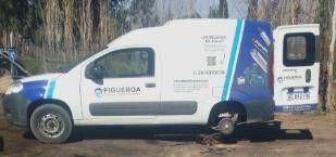 Una furgoneta comercial blanca y azul, a la que le falta una rueda trasera, estaba aparcada en un solar de tierra con árboles al fondo.