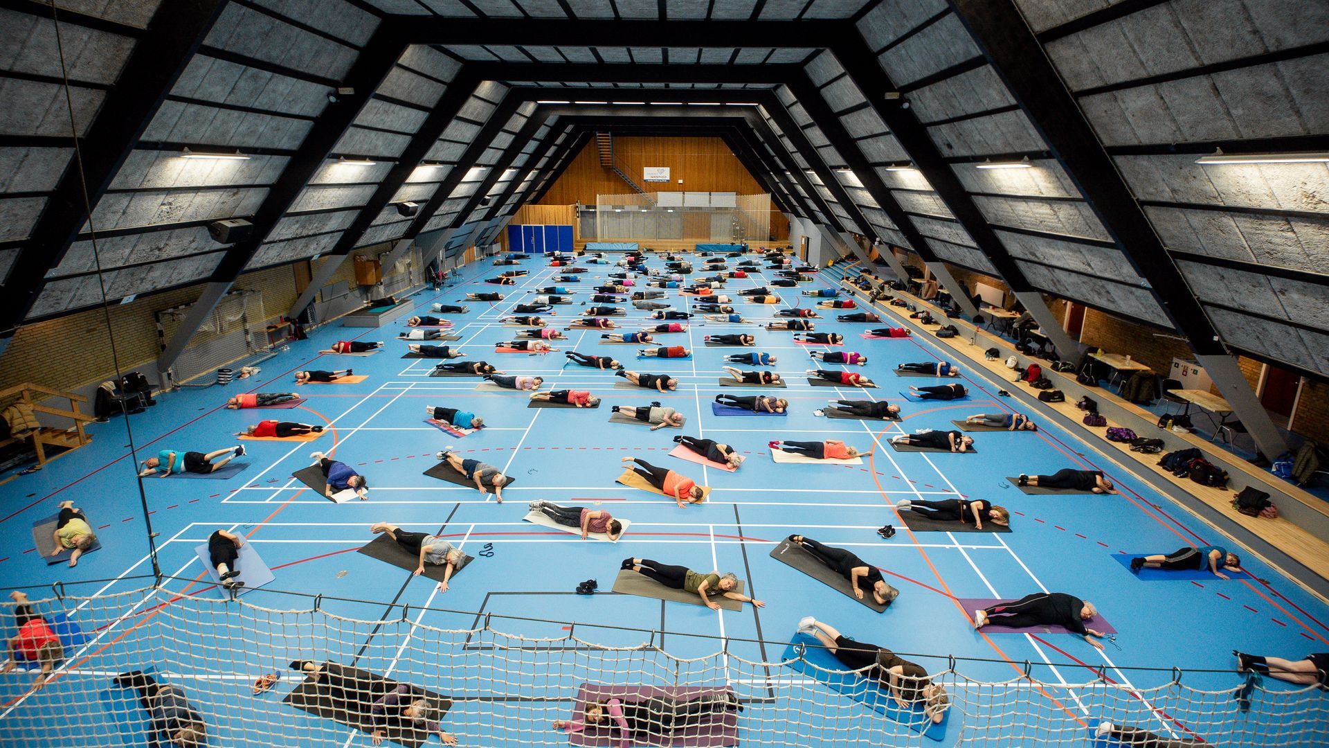 Stort indendørs fitnesscenter, mange mennesker på måtter i plankestillinger, under skrå loft. Blåt gulv, hvide linjer.