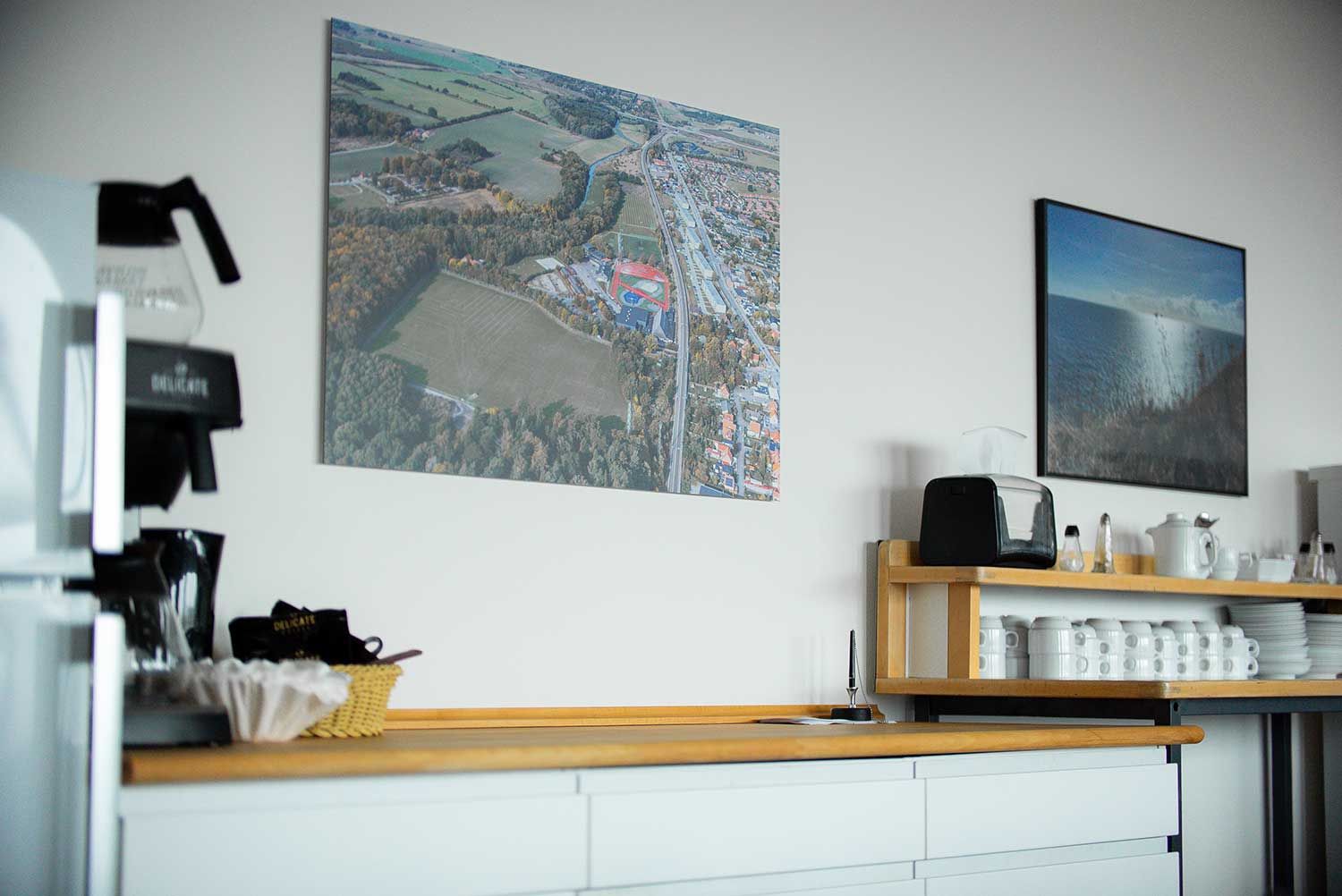 Køkken med indrammede landskabsfotos, kaffemaskine og bordplade med hvide skabe.