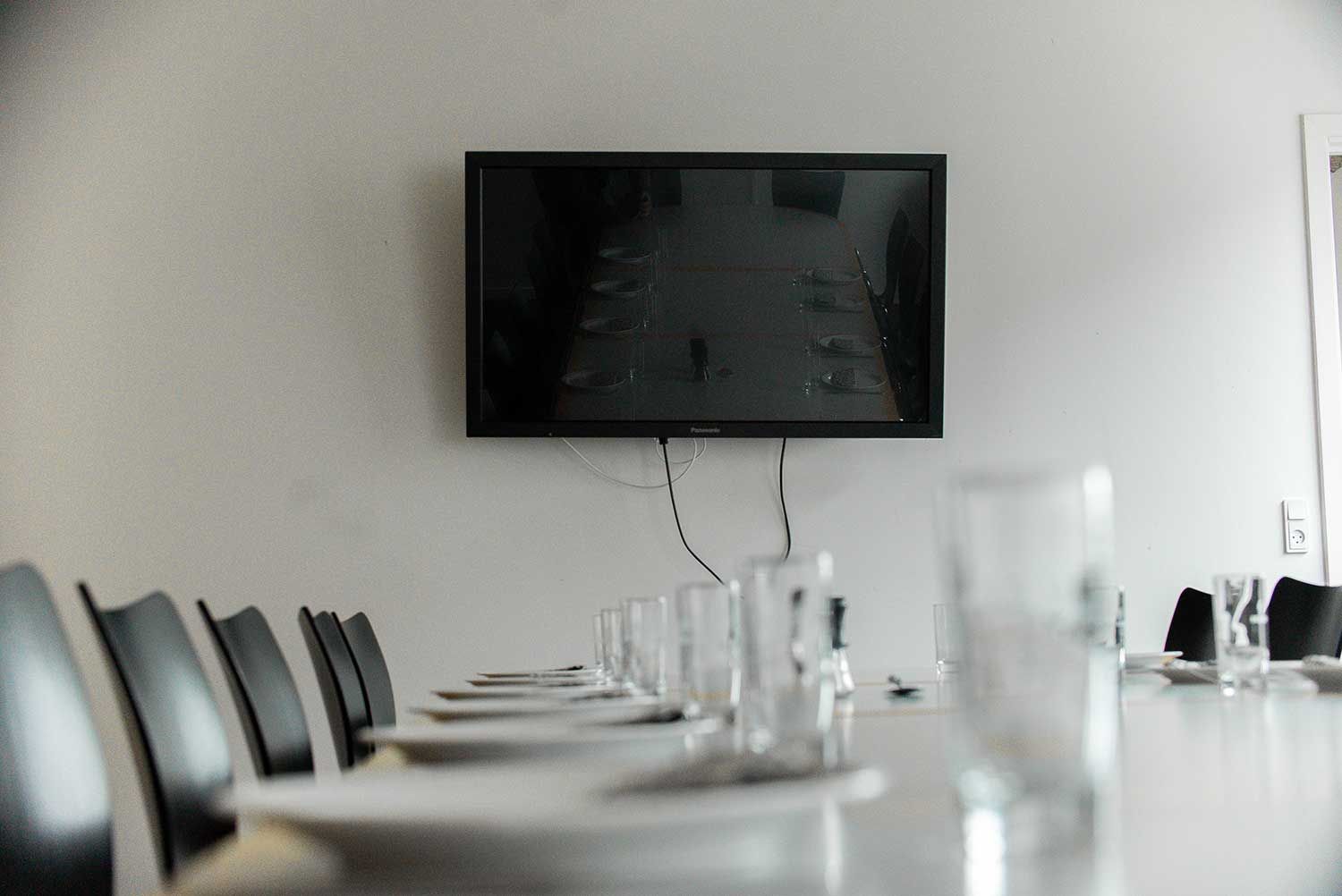 Et konferencerum med et vægmonteret TV og et borddækning med tallerkener og glas.