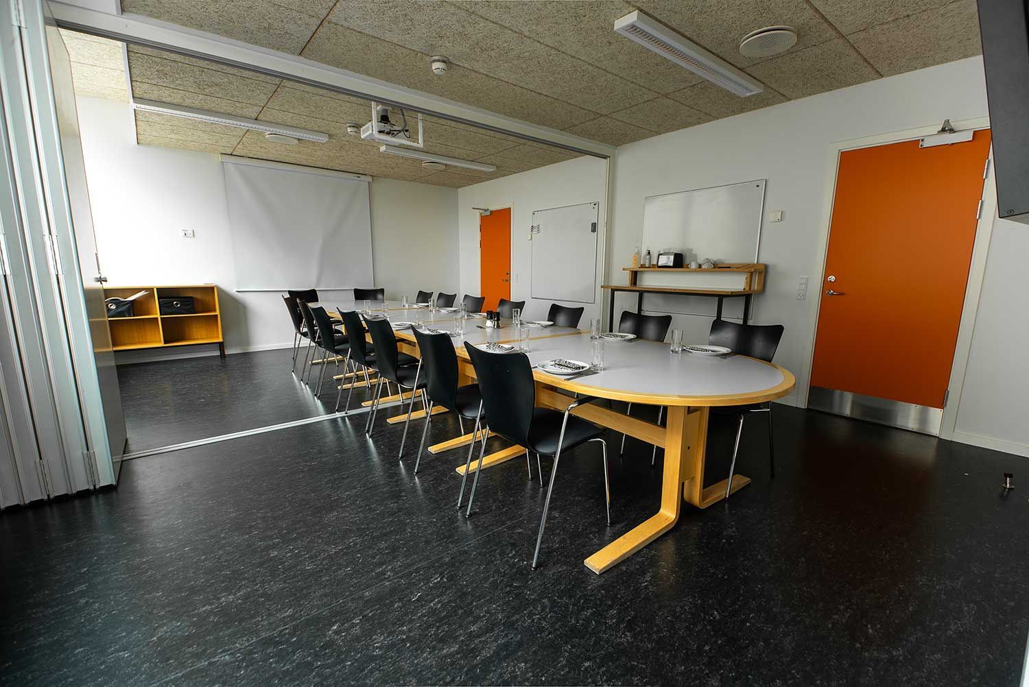 Konferencerum med ovalt bord, sorte stole, orange dør og projektorlærred.