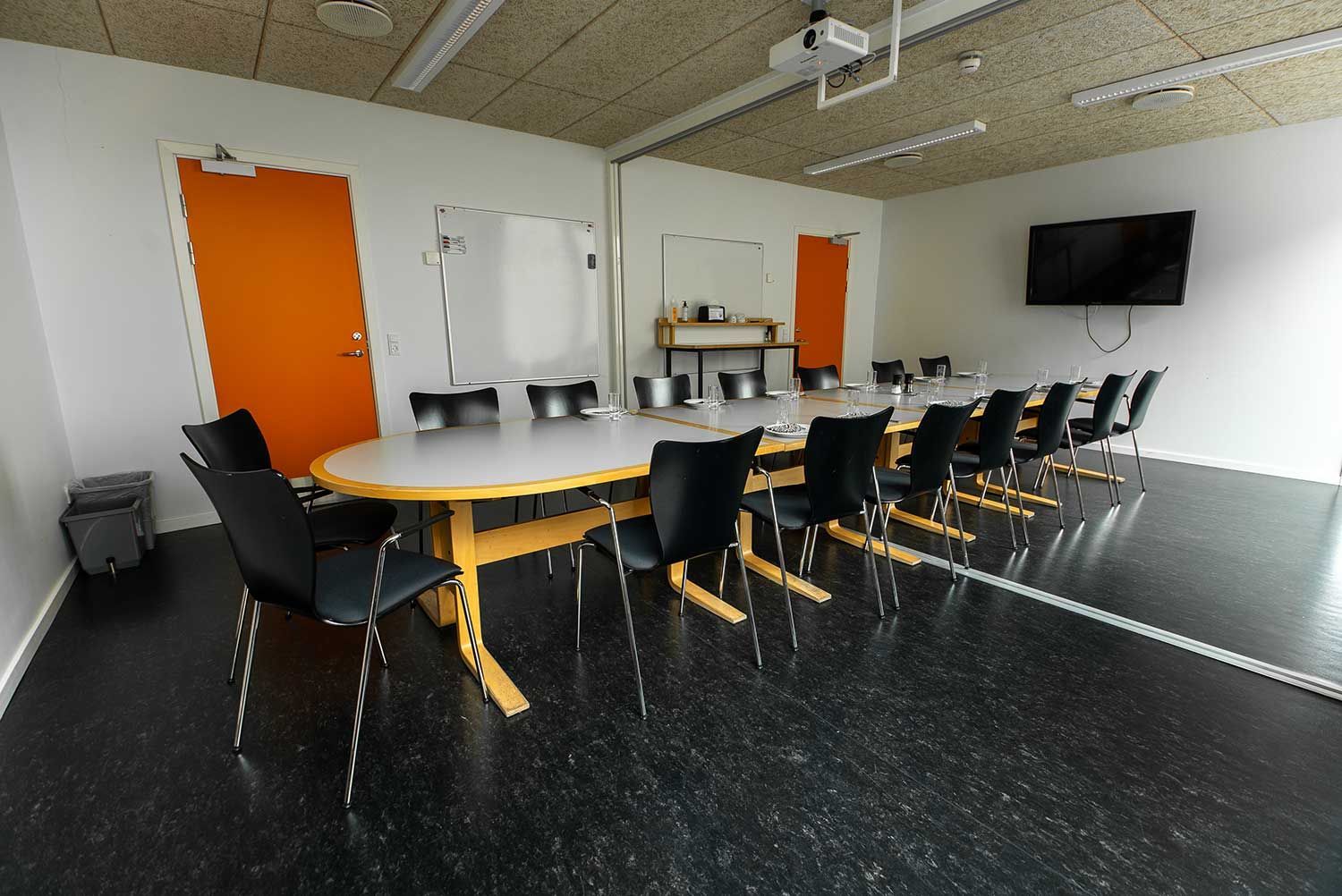 Konferencerum med langt bord, sorte stole, orange dør, whiteboards og TV.