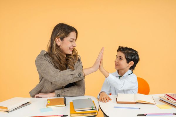 Profesora ayudando a un alumno a escribir en un pupitre cerca de unas ventanas luminosas.