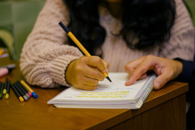 Una persona escribe en un cuaderno con un lápiz sobre una mesa de madera, con lápices de colores cerca.