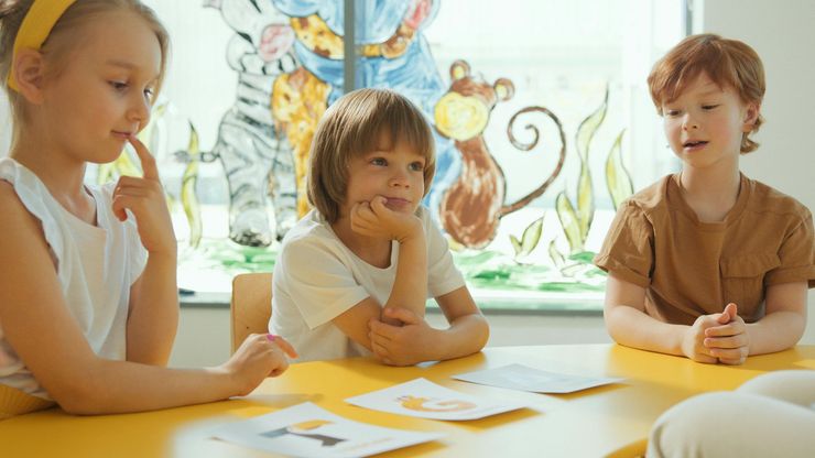 Tres niños sentados a una mesa con papeles, con expresión pensativa, en un aula luminosa.