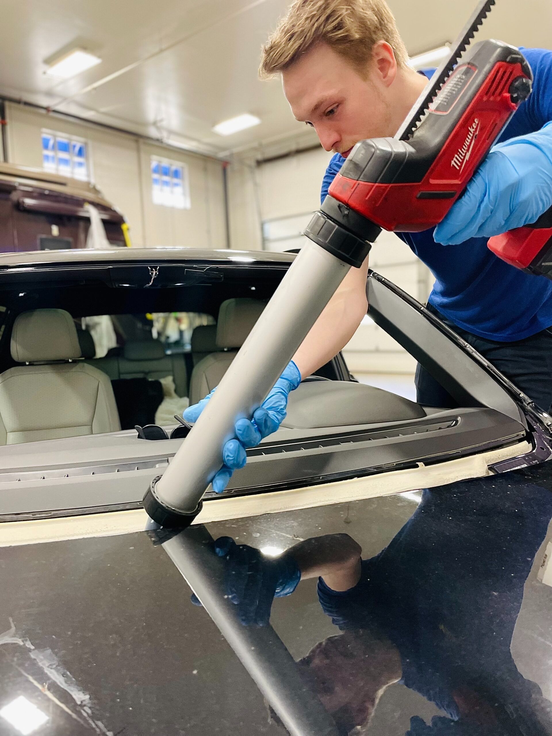 Person using a caulking gun to apply sealant to a car windshield in a garage.