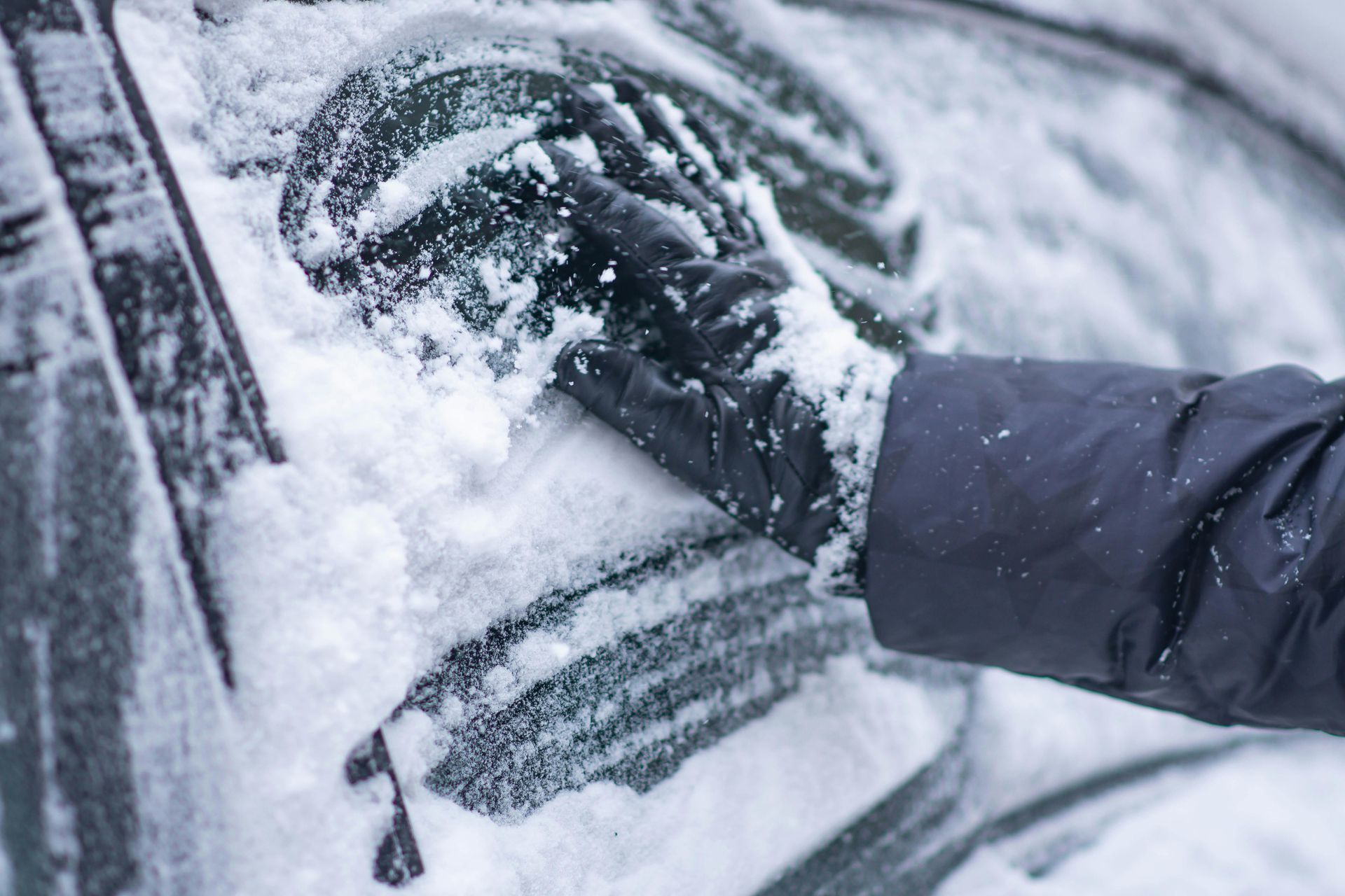 Gloved hand brushing snow from a car window in a winter setting.