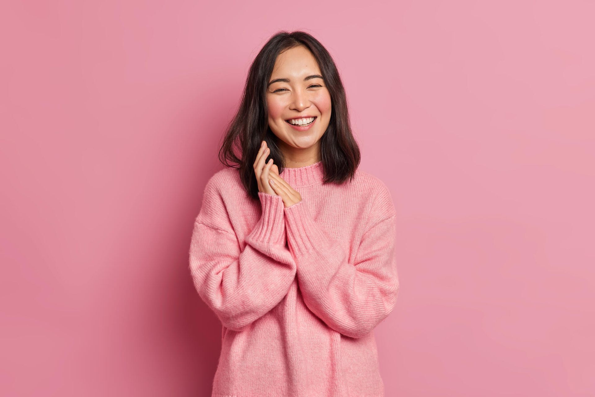 Woman in pink sweater laughs, hands clasped, in front of a pink background.