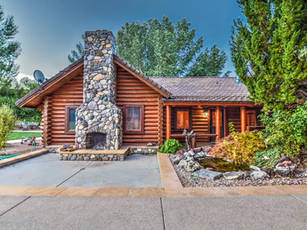 Log cabin with a stone chimney, porch, and small pond in a landscaped setting.