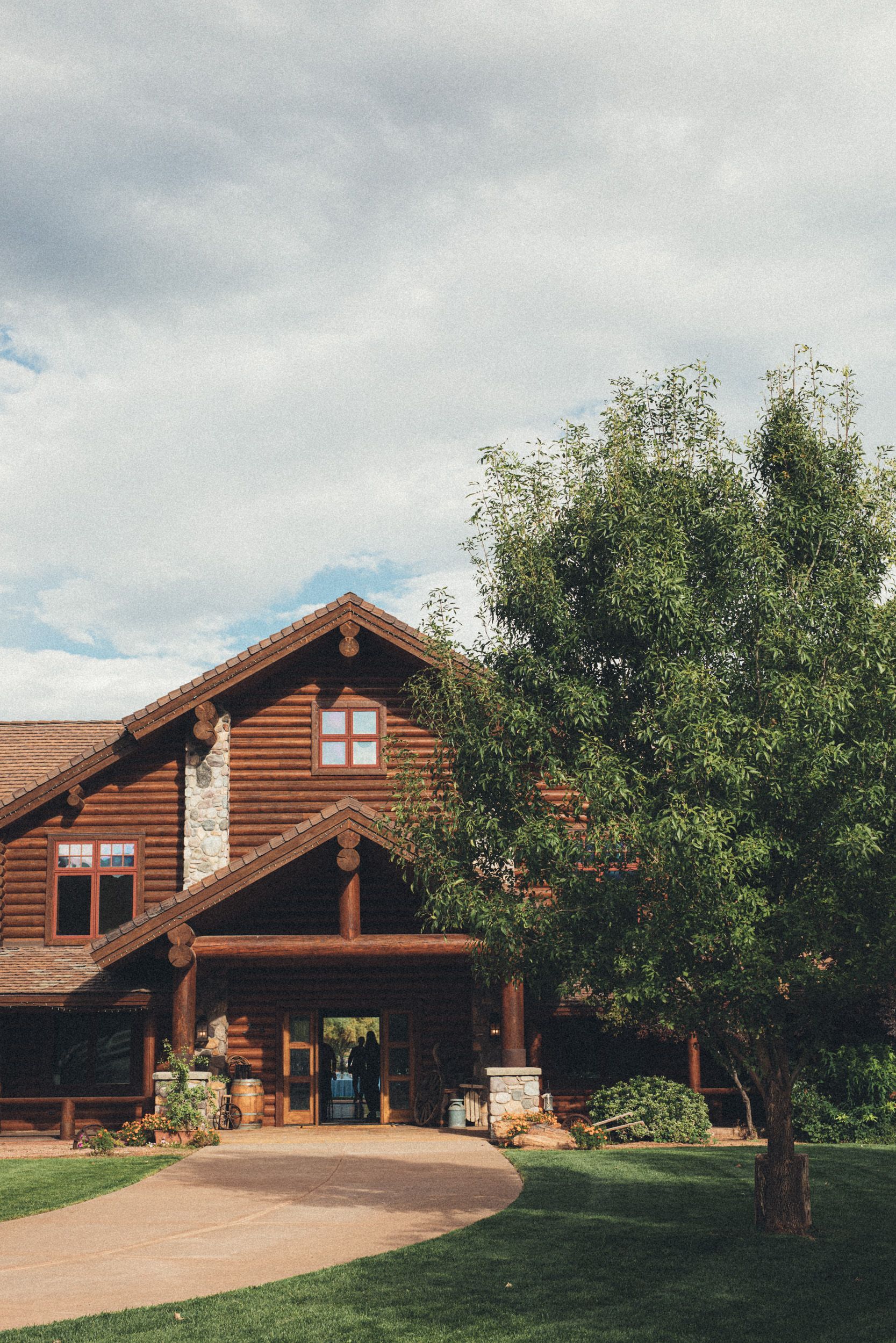 Log cabin with stone chimney, tree, and walkway under cloudy sky.