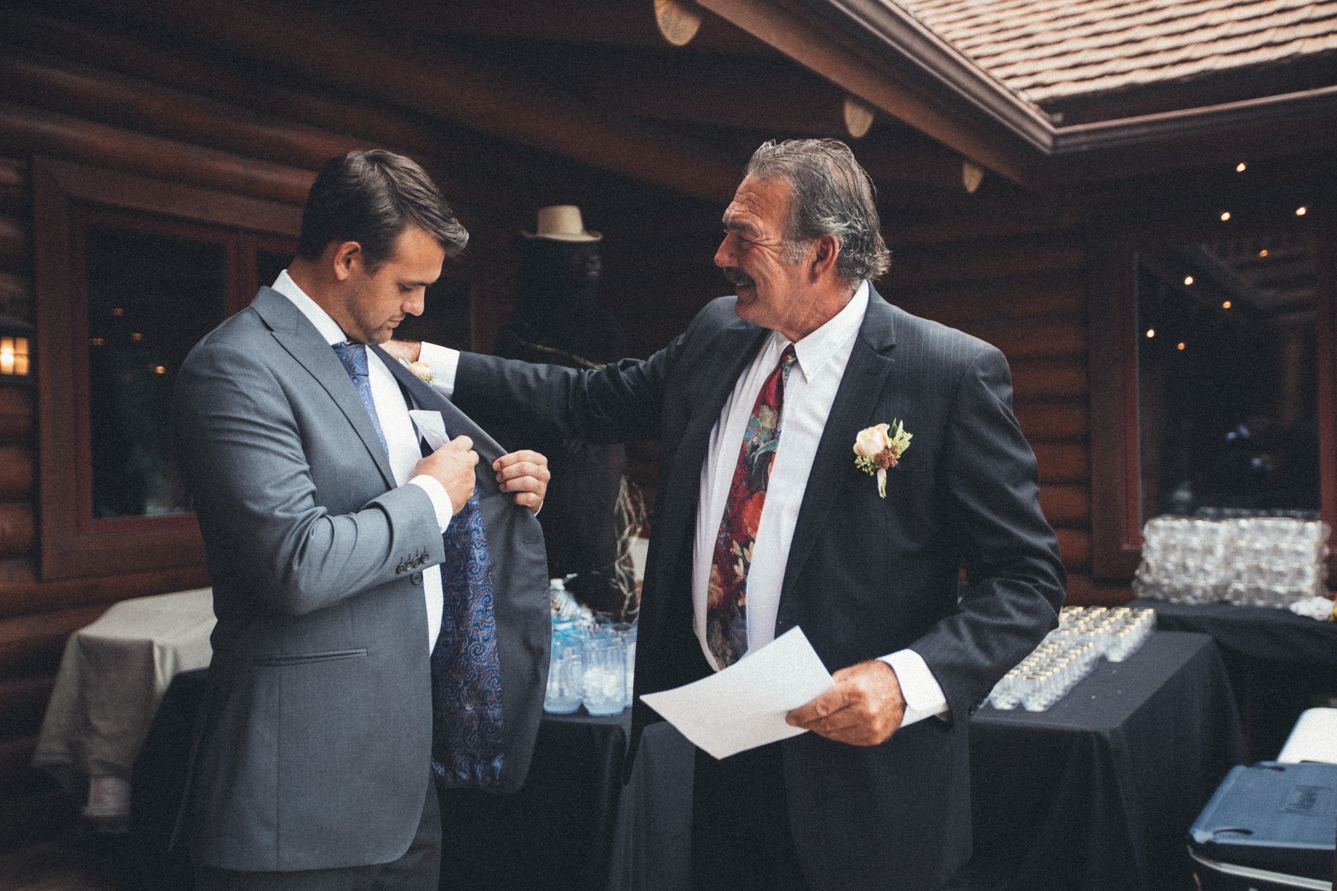 Man in suit adjusting another man's jacket pocket at an outdoor event. A table with drinks is in the background.