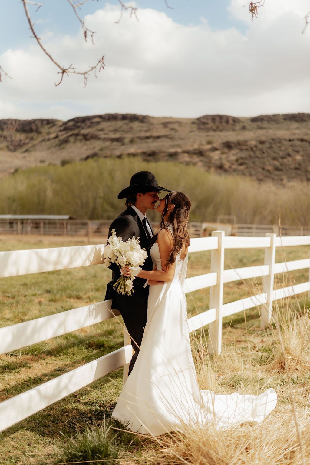 Log cabin with stone chimney, porch, and rocking chairs, set in a mountain landscape with trees and a snow-capped peak.