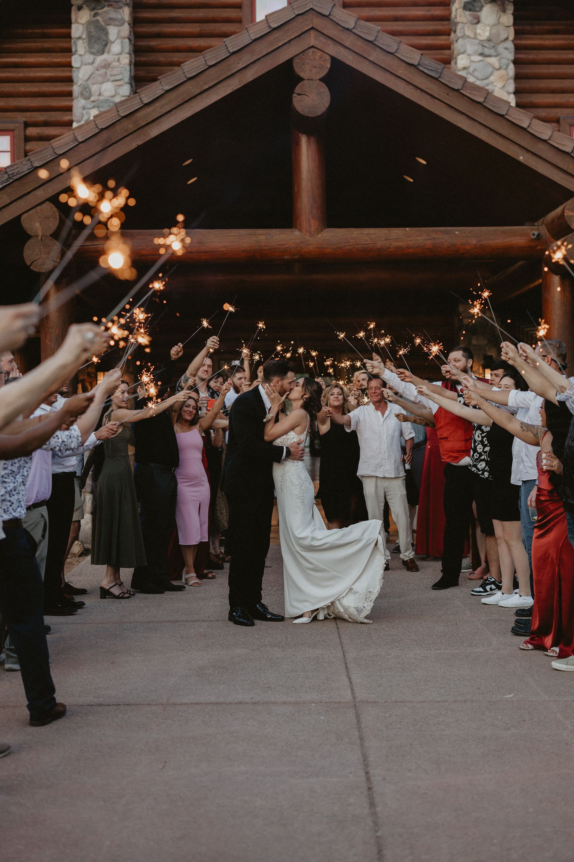 Couple kisses under sparklers held by guests at a wedding ceremony outside a wooden building.