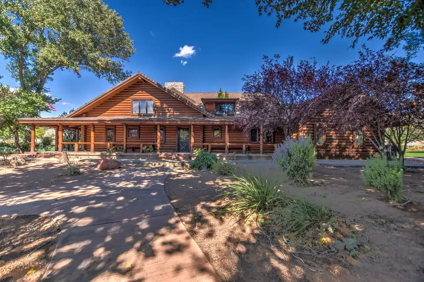 Log cabin home with covered porch, surrounded by trees and landscaping under a blue sky.