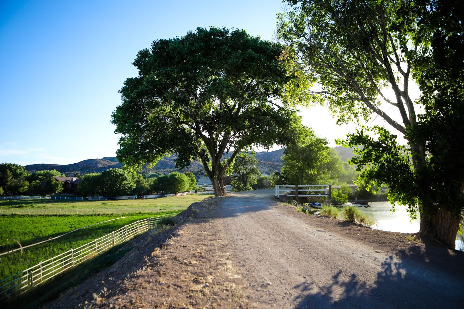 Grassy field with wooden fence, red rock mountains, and buildings under a clear blue sky.