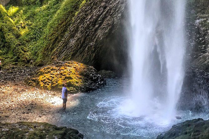 A person stands close to the base of a large waterfall, likely in the Columbia River Gorge near Portland, Oregon, surrounded by moss-covered rocks and mist from the waterfall.