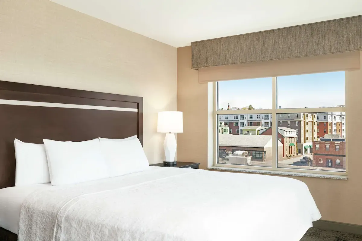 A bright and modern hotel room in Portland, Oregon, with a comfortable white bed and a window offering a view of downtown buildings, highlighting the urban landscape.