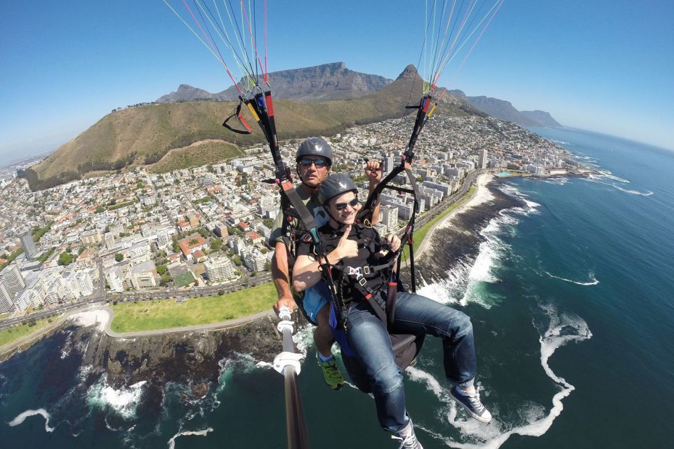 Two people tandem paragliding over a coastal city with mountains and the ocean visible below. They are flying high with parachute strings visible, capturing an exhilarating moment.